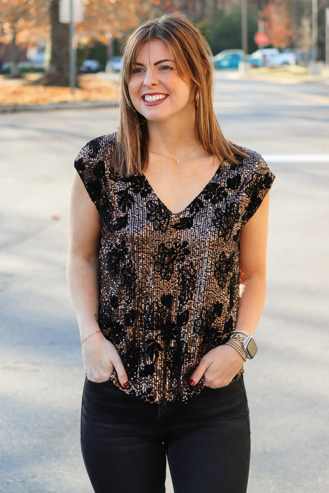 A brunette woman standing outside wearing a cap sleeve rose gold and black sequin top with a v neck and black jeans.
