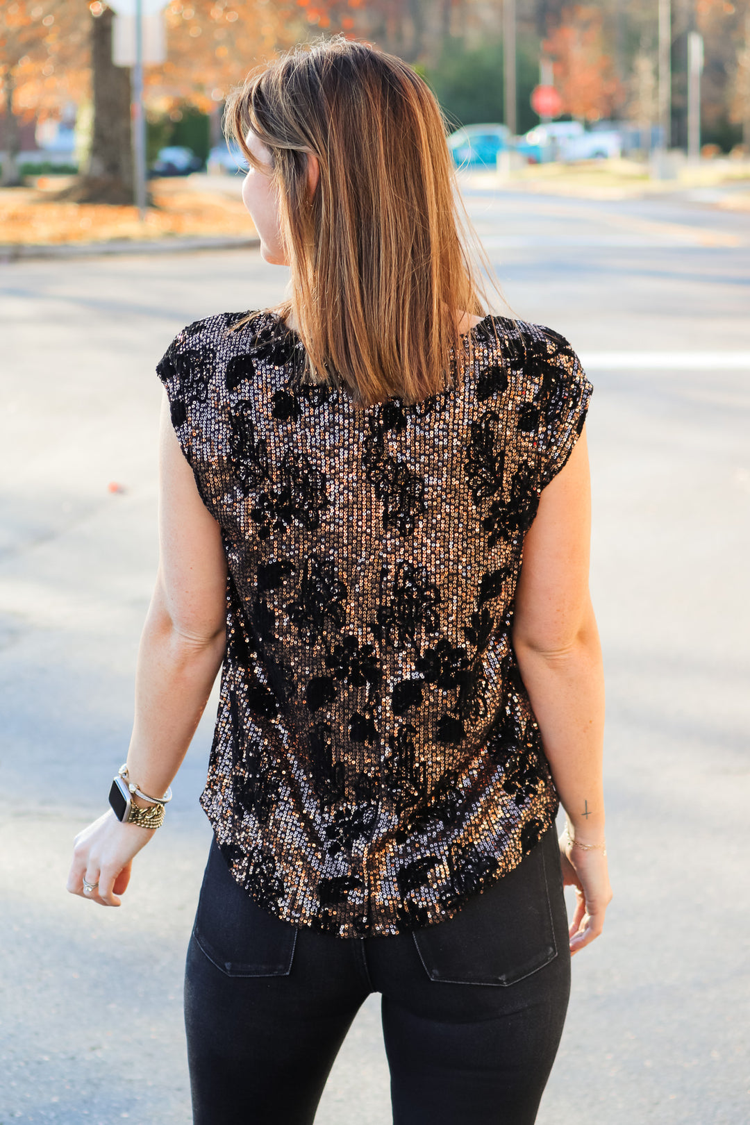 A brunette woman standing outside wearing a cap sleeve rose gold and black sequin top with a v neck and black jeans. She is rear facing.