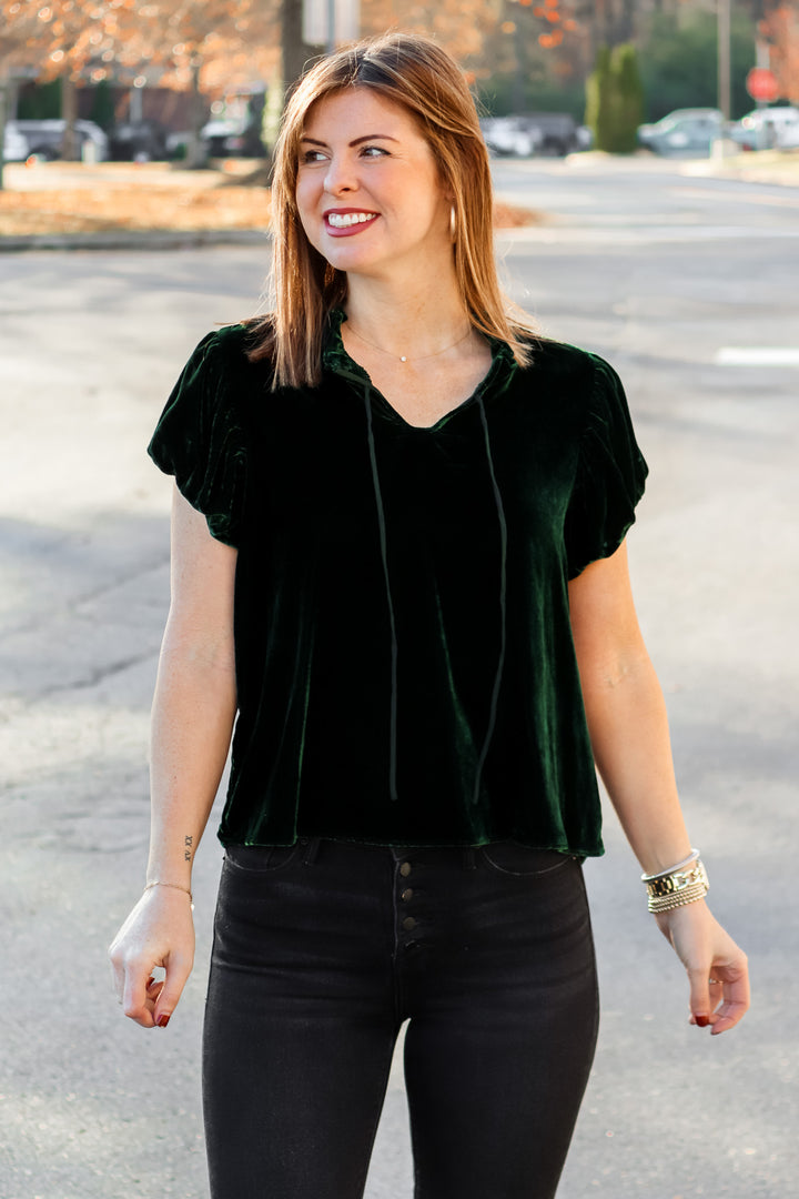 A brunette woman standing outside wearing a dark green velvet top with tie collar, short sleeves and black jeans.