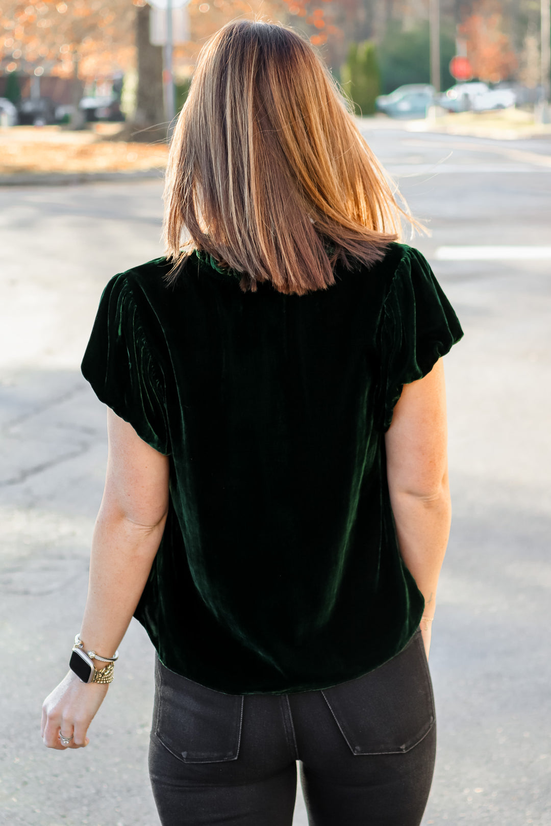 A brunette woman standing outside wearing a dark green velvet top with tie collar, short sleeves and black jeans. She is rear facing.
