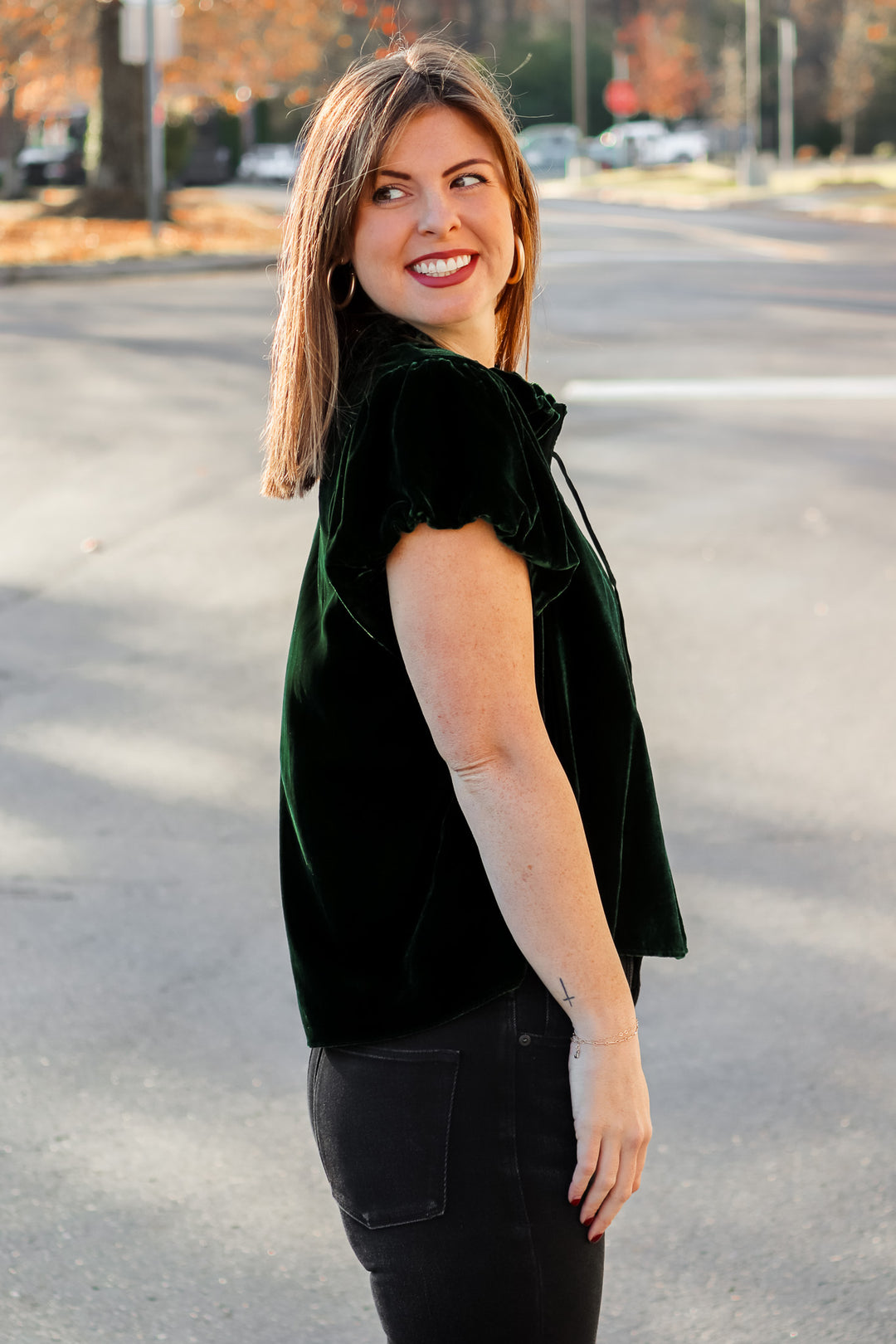 A brunette woman standing outside wearing a dark green velvet top with tie collar, short sleeves and black jeans.
