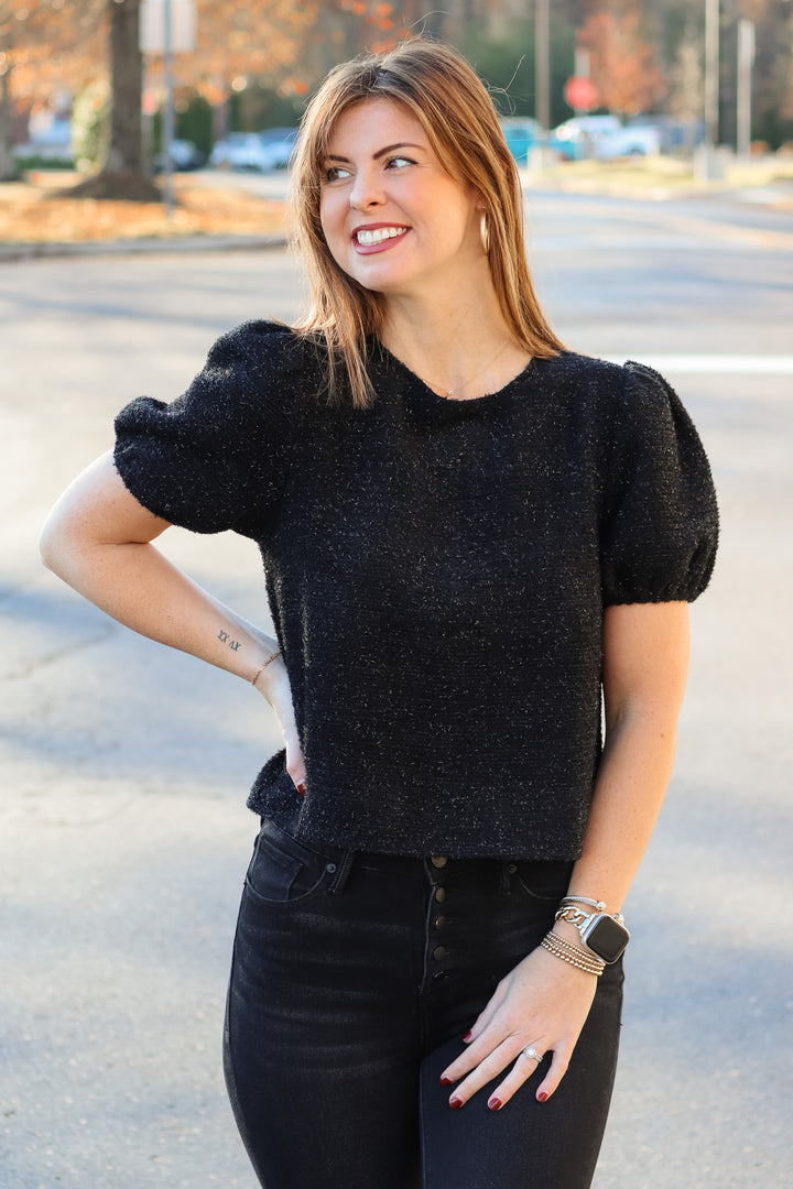 A brunette woman standing outside wearing a soft and shiny black top with short bubble sleeves and black jeans.