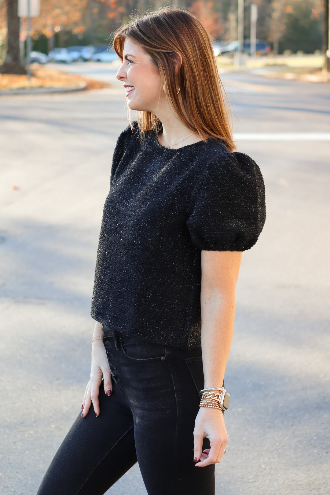 A brunette woman standing outside wearing a soft and shiny black top with short bubble sleeves and black jeans.