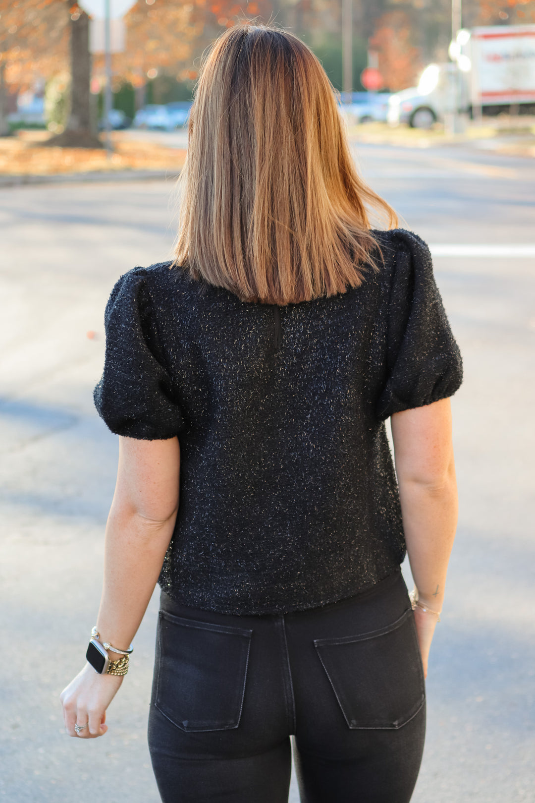 A brunette woman standing outside wearing a soft and shiny black top with short bubble sleeves and black jeans. She is rear facing.