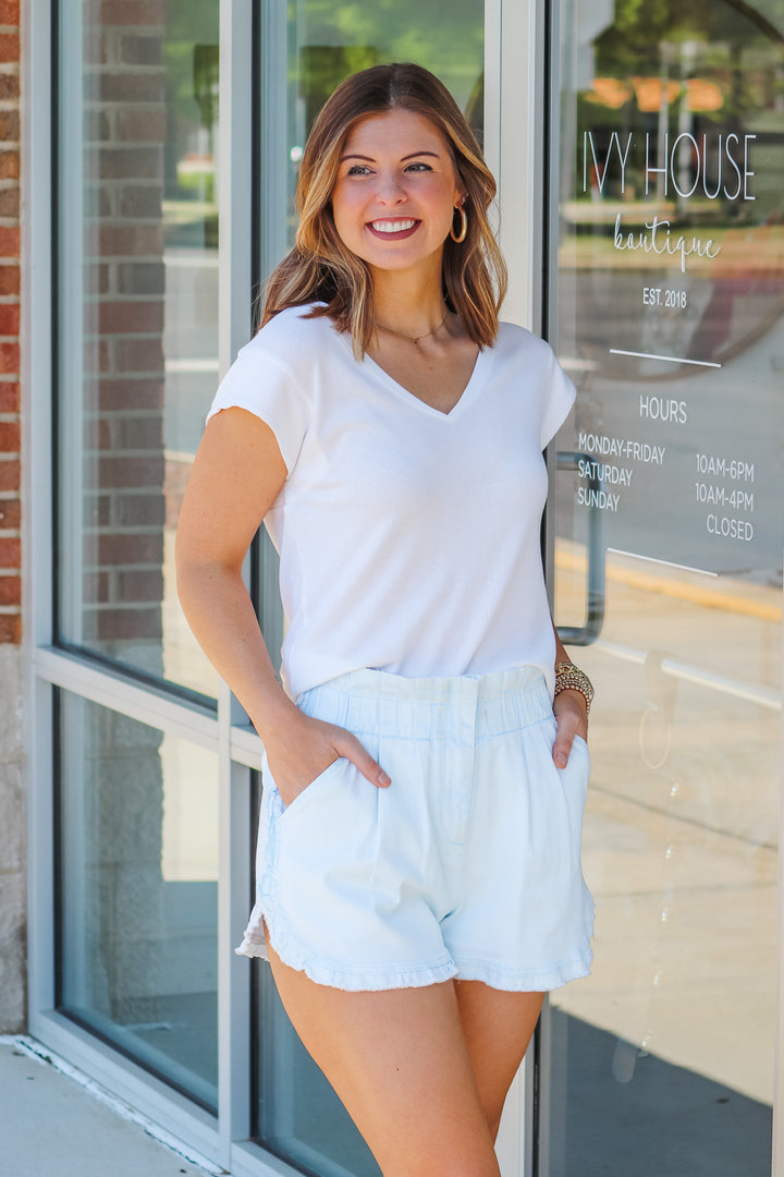 A brunette woman wearing a white shirt with cap sleeves and a v neck. She is standing in front of a shop.