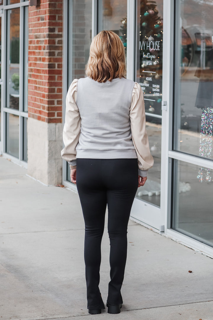 A woman standing in front of a shop wearing black front slit hem leggings and a neutral top. She is rear facing.